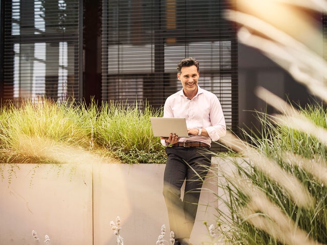 The image depicts a professional man standing outdoors, next to a modern building with large windows. He is smiling and holding an open laptop in his hands, appearing engaged and approachable. The man is dressed in business casual attire, wearing a light pink button-down shirt and dark trousers. He is positioned in front of a concrete planter filled with tall, green grasses, which adds a touch of nature to the urban setting. Sunlight filters through the plants, casting a warm, pleasant glow over the scene, contributing to an overall atmosphere of productivity and positivity.