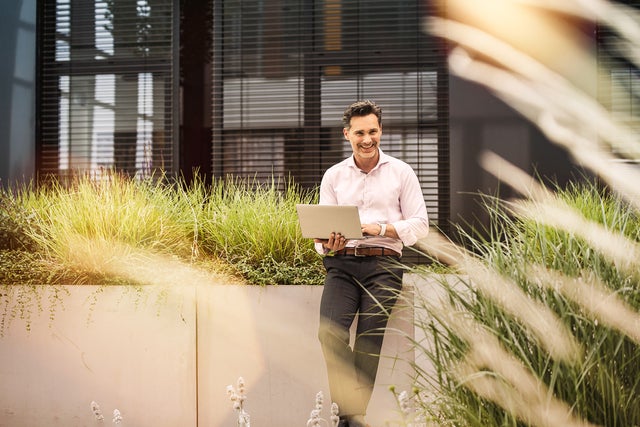 The image depicts a professional man standing outdoors, next to a modern building with large windows. He is smiling and holding an open laptop in his hands, appearing engaged and approachable. The man is dressed in business casual attire, wearing a light pink button-down shirt and dark trousers. He is positioned in front of a concrete planter filled with tall, green grasses, which adds a touch of nature to the urban setting. Sunlight filters through the plants, casting a warm, pleasant glow over the scene, contributing to an overall atmosphere of productivity and positivity.