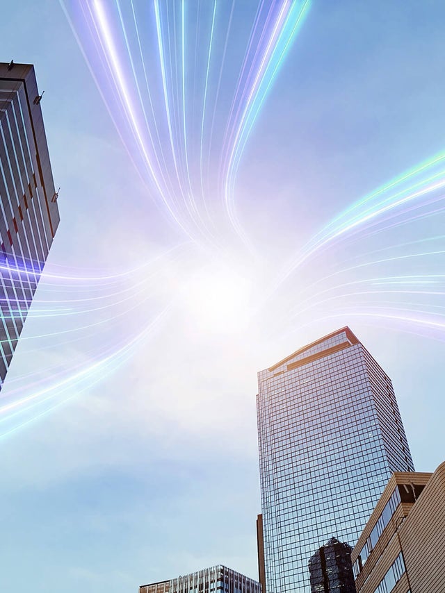 The image showcases a dynamic urban scene featuring several modern skyscrapers reaching towards the sky. The buildings are a mix of glass and steel structures, reflecting a professional and corporate environment. Superimposed over the scene are vibrant, multicolored light streaks that converge at a bright point in the sky, symbolizing connectivity and technology. The light streaks add a futuristic and energetic element to the image, suggesting advanced communication networks or data transfer within a bustling cityscape. The overall composition highlights the intersection of modern architecture and innovative technology.