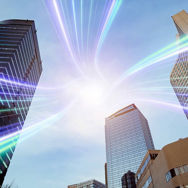 The image showcases a dynamic urban scene featuring several modern skyscrapers reaching towards the sky. The buildings are a mix of glass and steel structures, reflecting a professional and corporate environment. Superimposed over the scene are vibrant, multicolored light streaks that converge at a bright point in the sky, symbolizing connectivity and technology. The light streaks add a futuristic and energetic element to the image, suggesting advanced communication networks or data transfer within a bustling cityscape. The overall composition highlights the intersection of modern architecture and innovative technology.