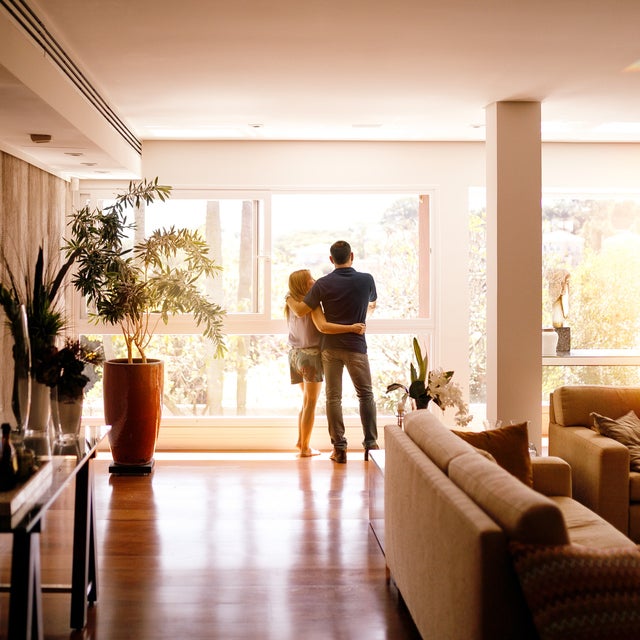 The picture shows a bright and spacious flat with large windows in the background that let in plenty of natural light. In the foreground, slightly shifted to the left, is a glass table with various decorative objects and bottles on it. On the left-hand side of the room is a large plant pot with a green plant that almost reaches the ceiling. In the centre of the picture, close to the large windows, a couple are standing closely together and looking outside. The man is wearing a dark blue shirt and jeans, while the woman is wearing a light-coloured top and shorts. They are standing on a shiny wooden floor that reflects the light. To the right of the picture is a beige sofa with several coloured cushions, which makes the living area cosy. Directly behind the sofa is a small table with other plants and decorations. The background shows a lush green landscape through the large windows, which enriches the room with a natural atmosphere.