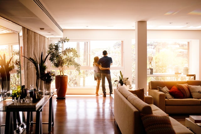 The picture shows a bright and spacious flat with large windows in the background that let in plenty of natural light. In the foreground, slightly shifted to the left, is a glass table with various decorative objects and bottles on it. On the left-hand side of the room is a large plant pot with a green plant that almost reaches the ceiling. In the centre of the picture, close to the large windows, a couple are standing closely together and looking outside. The man is wearing a dark blue shirt and jeans, while the woman is wearing a light-coloured top and shorts. They are standing on a shiny wooden floor that reflects the light. To the right of the picture is a beige sofa with several coloured cushions, which makes the living area cosy. Directly behind the sofa is a small table with other plants and decorations. The background shows a lush green landscape through the large windows, which enriches the room with a natural atmosphere.