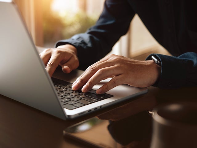 L'image montre un gros plan du clavier d'un ordinateur portable, qui est éclairé par des rayons de soleil chauds provenant de l'arrière-plan. L'écran de l'ordinateur portable se trouve sur le bord gauche de l'image et l'on peut voir une femme vêtue d'une chemise bleu foncé, assise dans un flou sur le bord droit. Ses mains reposent légèrement sur les touches de l'ordinateur portable. L'arrière-plan de l'image est très flou, mais on peut facilement reconnaître les murs d'une pièce et une fenêtre à travers laquelle passent les rayons du soleil.