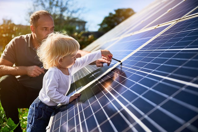 A father and his young child are engaging with solar panels outdoors. The father, kneeling beside the child, observes attentively while the child, with blonde hair, wearing a white shirt and blue jeans, uses a small tool to interact with the solar panel surface. The sunlight illuminates the scene, emphasizing the importance of renewable energy and family involvement in sustainable practices. This image highlights the hands-on approach to solar power management, symbolizing a commitment to future generations and environmentally responsible energy solutions.