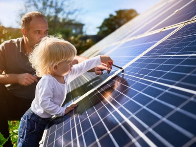 A father and his young child are engaging with solar panels outdoors. The father, kneeling beside the child, observes attentively while the child, with blonde hair, wearing a white shirt and blue jeans, uses a small tool to interact with the solar panel surface. The sunlight illuminates the scene, emphasizing the importance of renewable energy and family involvement in sustainable practices. This image highlights the hands-on approach to solar power management, symbolizing a commitment to future generations and environmentally responsible energy solutions.