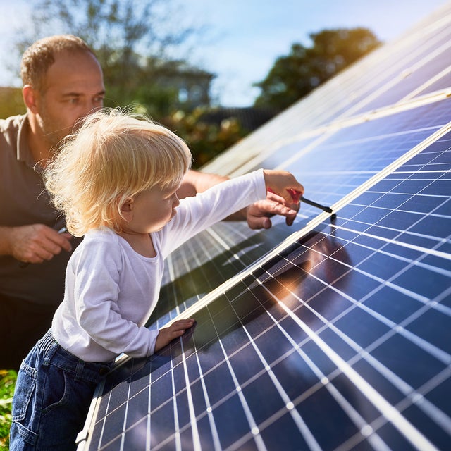 A father and his young child are engaging with solar panels outdoors. The father, kneeling beside the child, observes attentively while the child, with blonde hair, wearing a white shirt and blue jeans, uses a small tool to interact with the solar panel surface. The sunlight illuminates the scene, emphasizing the importance of renewable energy and family involvement in sustainable practices. This image highlights the hands-on approach to solar power management, symbolizing a commitment to future generations and environmentally responsible energy solutions.