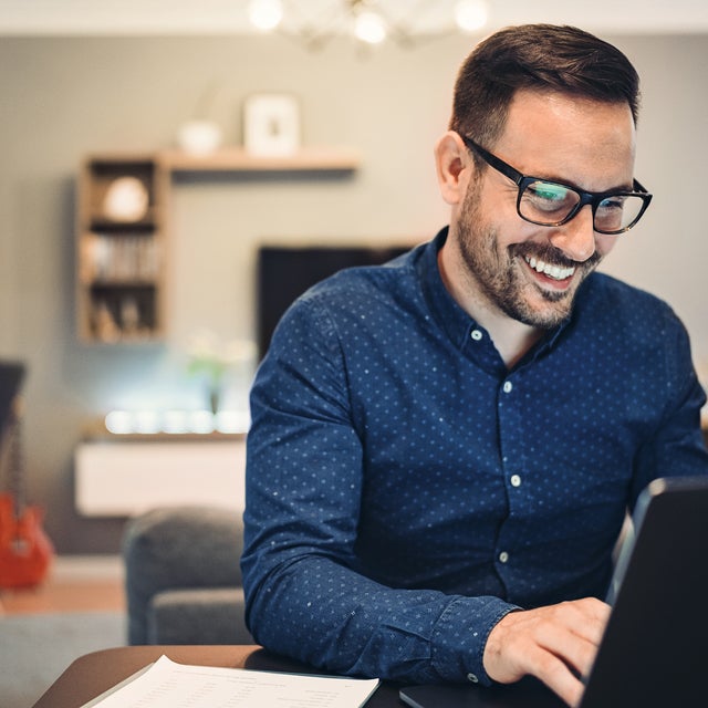 The image depicts a man sitting at a round table in a well-lit office space. There is a glass of water and a small espresso cup on the table, adding to the professional yet comfortable atmosphere. The man is smiling and engaged with the water consumption billing from ista on his laptop, taking notes in a notebook. He is wearing a blue button-down shirt and glasses. The background features a flip chart, potted plants, modern furniture, and large windows allowing natural light to flood the room. 