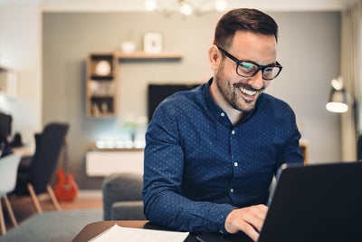 The image depicts a man sitting at a round table in a well-lit office space. There is a glass of water and a small espresso cup on the table, adding to the professional yet comfortable atmosphere. The man is smiling and engaged with the water consumption billing from ista on his laptop, taking notes in a notebook. He is wearing a blue button-down shirt and glasses. The background features a flip chart, potted plants, modern furniture, and large windows allowing natural light to flood the room. 