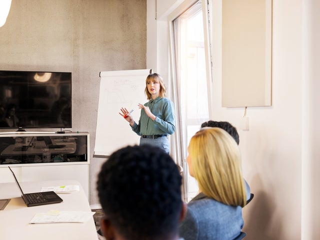 The image shows a business presentation in a conference room. A woman, standing at the front of the room, is presenting to a group of colleagues. She is wearing a light blue blouse and gray pants and is gesturing with a red marker in her hand. Behind her is a flip chart with a graph and some notes drawn on it. To her left, there is a large flat-screen monitor mounted on the wall. The attendees, who are seated and facing the presenter, are listening attentively. They include a mix of genders and ethnicities, dressed in professional attire. On the table in front of them are laptops, documents, and notepads, suggesting they are engaged in a formal business meeting. The room is well-lit, with a large window allowing natural light to enter, creating a bright and conducive environment for the presentation.