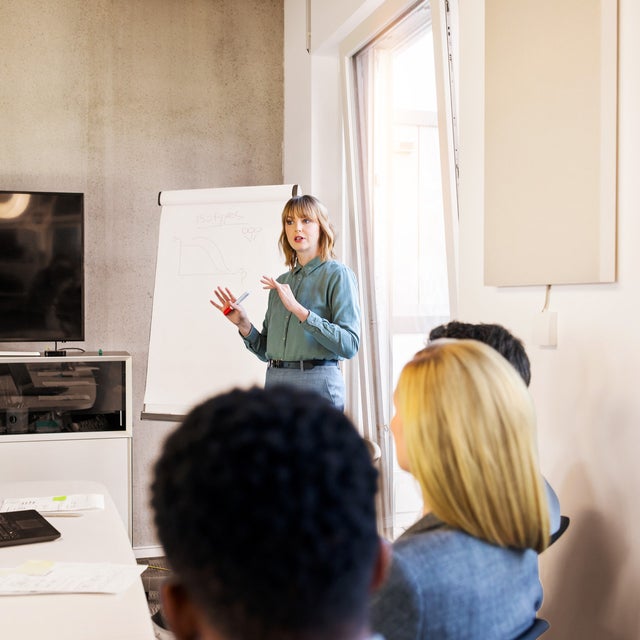 The image shows a business presentation in a conference room. A woman, standing at the front of the room, is presenting to a group of colleagues. She is wearing a light blue blouse and gray pants and is gesturing with a red marker in her hand. Behind her is a flip chart with a graph and some notes drawn on it. To her left, there is a large flat-screen monitor mounted on the wall. The attendees, who are seated and facing the presenter, are listening attentively. They include a mix of genders and ethnicities, dressed in professional attire. On the table in front of them are laptops, documents, and notepads, suggesting they are engaged in a formal business meeting. The room is well-lit, with a large window allowing natural light to enter, creating a bright and conducive environment for the presentation.