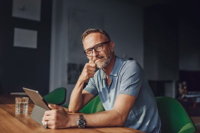 The picture shows a rather dark room that is barely lit with grey walls and a white partition. The only light only illuminates the dark wooden table at which a man is sitting in the foreground. The man sits at a green fabric chair with a short backrest and looks directly into the camera. The man has short light brown hair, a greying beard, wears black glasses, a light blue polo shirt and a large black watch on his left wrist. In front of him on the table is a glass of water and in his hand in front of him is a tablet on which he is informing himself about istas water-metering.