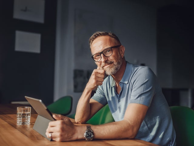 The picture shows a rather dark room that is barely lit with grey walls and a white partition. The only light only illuminates the dark wooden table at which a man is sitting in the foreground. The man sits at a green fabric chair with a short backrest and looks directly into the camera. The man has short light brown hair, a greying beard, wears black glasses, a light blue polo shirt and a large black watch on his left wrist. In front of him on the table is a glass of water and in his hand in front of him is a tablet on which he is informing himself about istas water-metering.