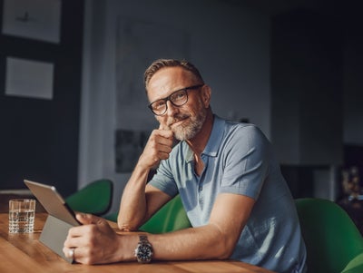 The picture shows a rather dark room that is barely lit with grey walls and a white partition. The only light only illuminates the dark wooden table at which a man is sitting in the foreground. The man sits at a green fabric chair with a short backrest and looks directly into the camera. The man has short light brown hair, a greying beard, wears black glasses, a light blue polo shirt and a large black watch on his left wrist. In front of him on the table is a glass of water and in his hand in front of him is a tablet on which he is informing himself about istas water-metering.