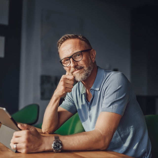 The picture shows a rather dark room that is barely lit with grey walls and a white partition. The only light only illuminates the dark wooden table at which a man is sitting in the foreground. The man sits at a green fabric chair with a short backrest and looks directly into the camera. The man has short light brown hair, a greying beard, wears black glasses, a light blue polo shirt and a large black watch on his left wrist. In front of him on the table is a glass of water and in his hand in front of him is a tablet on which he is informing himself about istas water-metering.