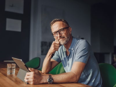 The picture shows a rather dark room that is barely lit with grey walls and a white partition. The only light only illuminates the dark wooden table at which a man is sitting in the foreground. The man sits at a green fabric chair with a short backrest and looks directly into the camera. The man has short light brown hair, a greying beard, wears black glasses, a light blue polo shirt and a large black watch on his left wrist. In front of him on the table is a glass of water and in his hand in front of him is a tablet on which he is informing himself about istas water-metering.