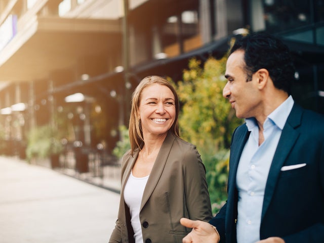 Two business professionals, a man and a woman, engage in a friendly conversation while walking outside near modern office buildings. The woman, dressed in a smart blazer, smiles warmly, while the man gestures as he speaks. The background features greenery and urban architecture, suggesting a productive, collaborative environment in a professional setting. The image reflects themes of business communication, networking, and positive workplace interactions.