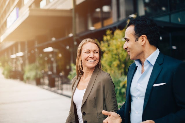 Two business professionals, a man and a woman, engage in a friendly conversation while walking outside near modern office buildings. The woman, dressed in a smart blazer, smiles warmly, while the man gestures as he speaks. The background features greenery and urban architecture, suggesting a productive, collaborative environment in a professional setting. The image reflects themes of business communication, networking, and positive workplace interactions.