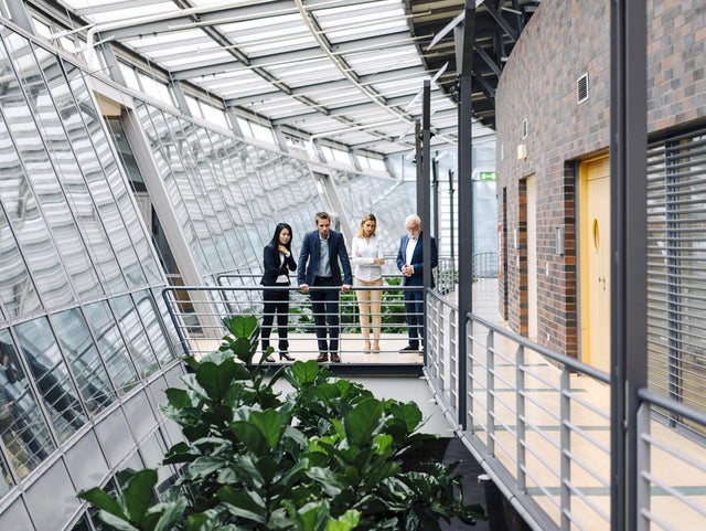 The picture shows four people in formal dress standing in a modern, light-flooded building. They are standing on a raised walkway with railings, surrounded by large glass windows. The windows extend along the entire length of the corridor and let in plenty of daylight.on the left of the picture is a woman with long, dark hair wearing a black trouser suit. She has her arms crossed in front of her chest and is looking down as if she is pensive. Next to her is a man in a dark blue suit, who is leaning on the railing with both hands and also looking down. Next to him is a woman with blonde hair in a white blazer and beige trousers. To her right is an older man with white hair and a beard, wearing a dark blue suit and interlocking fingers. The four people are discussing ista's building management system. Below the catwalk are large green plants that lend the room a natural element. On the right of the picture you can see a wall with dark red bricks and several doors. The whole space has a modern and open feel, with lots of natural light and a mix of industrial and natural elements.