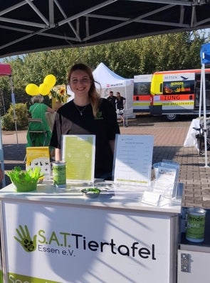 Eine junge Frau an einem Informationsstand der „S.A.T. Tiertafel Essen e.V.“ im Freien, unter einem Pavillon. Sie lächelt, trägt ein schwarzes T-Shirt mit ihrem Namensschild und ist von Informationsmaterialien und Spendenartikeln auf dem Tisch umgeben. Im Hintergrund sind weitere Stände und Personen zu sehen.