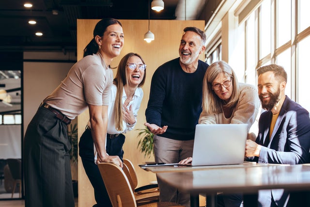 A diverse group of five professionals, spanning different ages, gather around a laptop in a modern office space. They are engaged in a lively discussion, with smiles and laughter, reflecting teamwork, collaboration, and a positive work environment. The sunlight streams through large windows, creating a warm, inviting atmosphere that highlights the dynamic of the team.