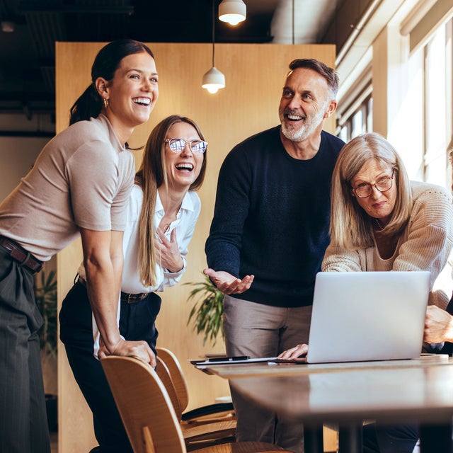 A diverse group of five professionals, spanning different ages, gather around a laptop in a modern office space. They are engaged in a lively discussion, with smiles and laughter, reflecting teamwork, collaboration, and a positive work environment. The sunlight streams through large windows, creating a warm, inviting atmosphere that highlights the dynamic of the team.