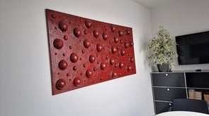 Ein rotes, strukturiertes Kunstwerk hängt an einer weißen Wand in einem Raum. Es zeigt viele dreidimensionale, runde Formen, die auf der Oberfläche hervortreten. Rechts steht eine Zimmerpflanze neben einem Fernseher.