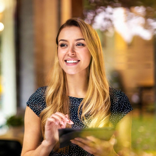 The picture shows a young woman looking through a window. She has long blonde hair and is wearing a dark blue, polka-dotted top. The woman is smiling and holding a tablet in both hands which is showing the Energy Audit Reporting from ista, her gaze directed slightly to the right. Green plants and a blurred light source can be seen in the background, giving the picture a warm and inviting atmosphere. The light source is located approximately in the centre of the background. The left background area shows a vertical black line, which is probably part of the window frame. The woman is standing relatively close to the window so that her reflection is partially visible in the glass. The foreground of the image is in focus while the background remains blurred, which draws the viewer's attention to the woman's smiling face and her tablet.