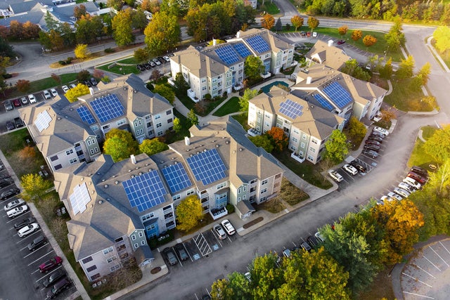 An aerial view of a residential complex showcasing several multi-story apartment buildings equipped with rooftop solar panels. The buildings are arranged in a circular layout, surrounded by well-maintained greenery, trees, and parking lots filled with vehicles. The roads curve around the complex, connecting the various structures. This image highlights the implementation of renewable energy through solar panels in a modern residential community, emphasizing sustainability and eco-friendly living practices. It illustrates the community's commitment to reducing its carbon footprint and embracing innovative energy solutions.