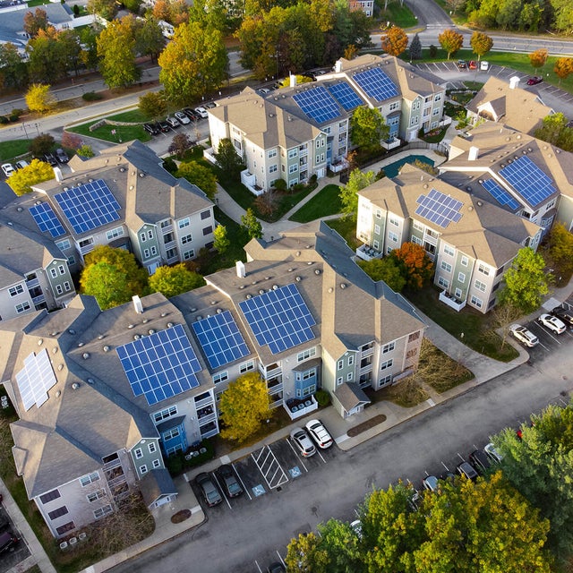 An aerial view of a residential complex showcasing several multi-story apartment buildings equipped with rooftop solar panels. The buildings are arranged in a circular layout, surrounded by well-maintained greenery, trees, and parking lots filled with vehicles. The roads curve around the complex, connecting the various structures. This image highlights the implementation of renewable energy through solar panels in a modern residential community, emphasizing sustainability and eco-friendly living practices. It illustrates the community's commitment to reducing its carbon footprint and embracing innovative energy solutions.