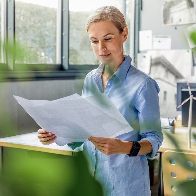 Durch die grünen Zweige einer Topfpflanze, sieht man eine Frau in einem Büro stehen. Sie steht mittig im Bild, trägt eine blaue Bluse und studiert Unterlagen, die Sie halbhoch in der Hand hält. Im Hintergrund ist ein Schreibtisch mit Bildschirm zu erkennen und eine Pinnwand.  