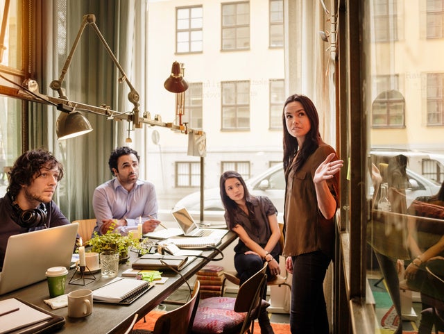 A team of four professionals is engaged in a meeting within a contemporary office space. A woman, standing and gesturing towards a large window, leads the discussion. Three colleagues—two men and one woman—sit attentively at a table equipped with laptops, notebooks, and coffee cups. The office is illuminated with natural light from expansive windows, offering a view of urban buildings outside. The setting reflects a collaborative and focused work environment, emphasizing the team's commitment to a culture of open communication, active participation, and collective problem-solving. This image underscores the importance of fostering a workplace culture that values teamwork and shared goals.