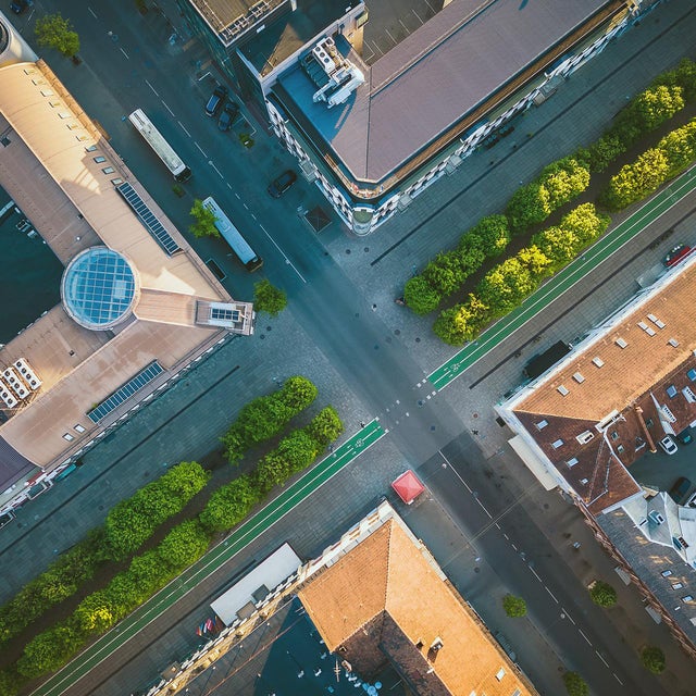 The picture shows a bird's eye view of the city. The picture was taken vertically downwards and shows an intersection from above, with two roads crossing in the centre of the picture. The road running from bottom left to top right is wider and appears to be a main road. It is lined with trees and a green strip on both sides that serves as a cycle path. The intersecting road, which runs from top left to bottom right, is slightly narrower and there are buildings with different roof shapes and colours at the four corners of the intersection. The building on the left has a striking, light brown roof with a round glass dome in the centre. Next to this building you can see a large bus parked on the street. The building at the top has a darker roof and is surrounded by several trees. The buildings on the right and below have red tiled roofs. The building on the right has a few cars in a car park next to it and the streets are quite clean with few vehicles or people to be seen. The green trees along the streets add a touch of nature to the urban environment. Overall, the picture appears calm and orderly, with clean lines and well-maintained urban structures.