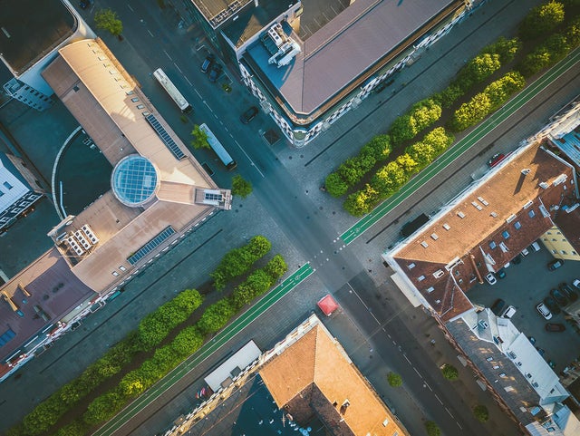 The picture shows a bird's eye view of the city. The picture was taken vertically downwards and shows an intersection from above, with two roads crossing in the centre of the picture. The road running from bottom left to top right is wider and appears to be a main road. It is lined with trees and a green strip on both sides that serves as a cycle path. The intersecting road, which runs from top left to bottom right, is slightly narrower and there are buildings with different roof shapes and colours at the four corners of the intersection. The building on the left has a striking, light brown roof with a round glass dome in the centre. Next to this building you can see a large bus parked on the street. The building at the top has a darker roof and is surrounded by several trees. The buildings on the right and below have red tiled roofs. The building on the right has a few cars in a car park next to it and the streets are quite clean with few vehicles or people to be seen. The green trees along the streets add a touch of nature to the urban environment. Overall, the picture appears calm and orderly, with clean lines and well-maintained urban structures.