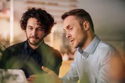 The picture shows two men looking intently at a laptop. The man on the left has curly, dark hair and a beard, while the man on the right has shorter, lighter-coloured hair. Both are wearing shirts, with the man on the left wearing a dark-coloured shirt and the man on the right a light-coloured shirt. They appear to be engaged in a conversation and are possibly working on a project together. There are blurred green leaves in the foreground, suggesting a leafy or natural environment. The background is blurred, but it can be recognised that they are in a modern office or workspace. You can dimly make out office furniture and shelving, suggesting a professional environment.