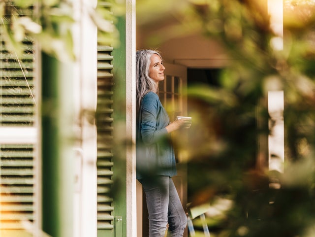 The picture shows a woman with grey, long hair who is the focus of the photo. She is wearing a blue cardigan and grey jeans. Her arms are bent in front of her body and she is holding a cup in her hands while leaning against a wall and looking to the right. To the left of the woman is a large green wooden grille, indicating a window. An open entrance door can be seen in the background on the right. Blurred green plants can be seen in the foreground on the right and top left, giving the picture a warm and cosy atmosphere.