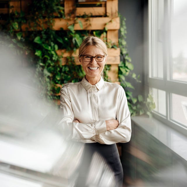 The picture shows a smiling woman with blonde hair and glasses standing in a modern office. She is wearing a cream-coloured blouse and has her arms crossed in front of her chest. The woman is in the centre of the picture. Behind her, a wall is covered with lush green foliage growing in a wooden frame. This green wall takes up the left part of the background and gives the room a natural and lively atmosphere. To the right of the woman, along the right side of the painting, is a window front that allows plenty of natural light into the room. The windows show a view of the roofs and the surroundings outside the building. A blur is visible in the foreground of the picture, possibly created by a glass surface or reflection. This blurring gives the image a certain depth and a modern photographic effect. The whole scene conveys an image of a modern, well-lit and green working environment that is both professional and inviting.