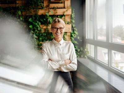 The picture shows a smiling woman with blonde hair and glasses standing in a modern office. She is wearing a cream-coloured blouse and has her arms crossed in front of her chest. The woman is in the centre of the picture. Behind her, a wall is covered with lush green foliage growing in a wooden frame. This green wall takes up the left part of the background and gives the room a natural and lively atmosphere. To the right of the woman, along the right side of the painting, is a window front that allows plenty of natural light into the room. The windows show a view of the roofs and the surroundings outside the building. A blur is visible in the foreground of the picture, possibly created by a glass surface or reflection. This blurring gives the image a certain depth and a modern photographic effect. The whole scene conveys an image of a modern, well-lit and green working environment that is both professional and inviting.