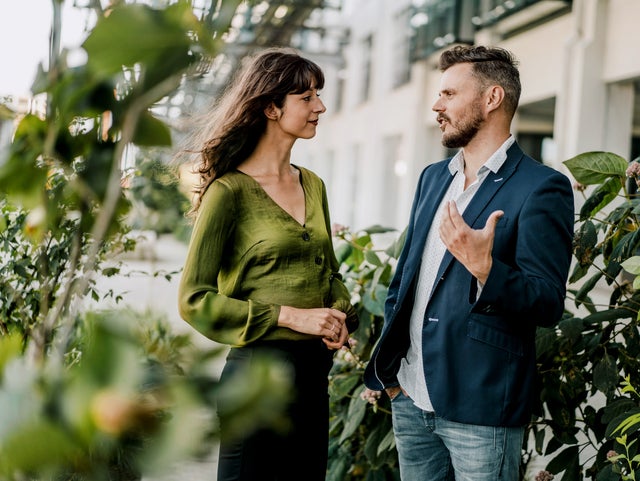 The picture shows a relaxed and friendly scene in which a woman and a man are chatting outdoors. The woman is on the left of the picture. She has long, slightly wavy dark hair and is wearing a green blouse with long sleeves and a V-neck. She holds her hands in front of her in a relaxed manner. The man on the right of the picture has short, dark hair. He is wearing a blue blazer over a white shirt and light-coloured jeans. As he speaks, he gestures with his right hand as if he is explaining or emphasising something. The background of the scene shows a modern building with large windows and light-coloured facades, suggesting an urban environment, possibly a terrace or courtyard. There are blurred green plants in the foreground and on the left-hand side of the picture, giving the image depth and creating a natural atmosphere. The light in the scene is natural and comes from the left side, which illuminates the scene well and creates a friendly, inviting mood. The overall atmosphere of the picture looks professional and at the same time relaxed, as if the two people are having a pleasant conversation. The combination of urban surroundings and nature conveys a sense of balance and harmony.