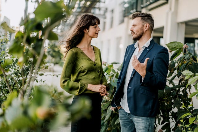 The picture shows a relaxed and friendly scene in which a woman and a man are chatting outdoors. The woman is on the left of the picture. She has long, slightly wavy dark hair and is wearing a green blouse with long sleeves and a V-neck. She holds her hands in front of her in a relaxed manner. The man on the right of the picture has short, dark hair. He is wearing a blue blazer over a white shirt and light-coloured jeans. As he speaks, he gestures with his right hand as if he is explaining or emphasising something. The background of the scene shows a modern building with large windows and light-coloured facades, suggesting an urban environment, possibly a terrace or courtyard. There are blurred green plants in the foreground and on the left-hand side of the picture, giving the image depth and creating a natural atmosphere. The light in the scene is natural and comes from the left side, which illuminates the scene well and creates a friendly, inviting mood. The overall atmosphere of the picture looks professional and at the same time relaxed, as if the two people are having a pleasant conversation. The combination of urban surroundings and nature conveys a sense of balance and harmony.