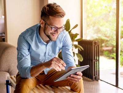 L'image montre un jeune homme à lunettes, légèrement penché vers l'avant et assis sur un banc moderne en bois. Le banc se trouve dans une pièce lumineuse et agréable. L'homme porte une chemise bleu clair et un pantalon couleur moutarde. Il tient une tablette dans ses mains et regarde le système de mesure de la chaleur d'ista. Derrière l'homme, à droite de l'image, se trouve une grande fenêtre allant du sol au plafond qui laisse entrer beaucoup de lumière naturelle dans la pièce. Un jardin verdoyant avec une végétation luxuriante est visible à travers la fenêtre. À côté de la fenêtre se trouve une grande plante dans un pot posé sur le sol. À droite de la plante se trouve un radiateur noir, qui contraste avec la lumière et les couleurs naturelles de la pièce. À gauche de l'homme, on aperçoit une partie d'un canapé beige. D'autres parties de la pièce sont visibles à l'arrière-plan, à gauche de l'image. Il peut s'agir d'une porte ou d'une autre fenêtre, qui apportent également de la lumière et de l'espace à la pièce.