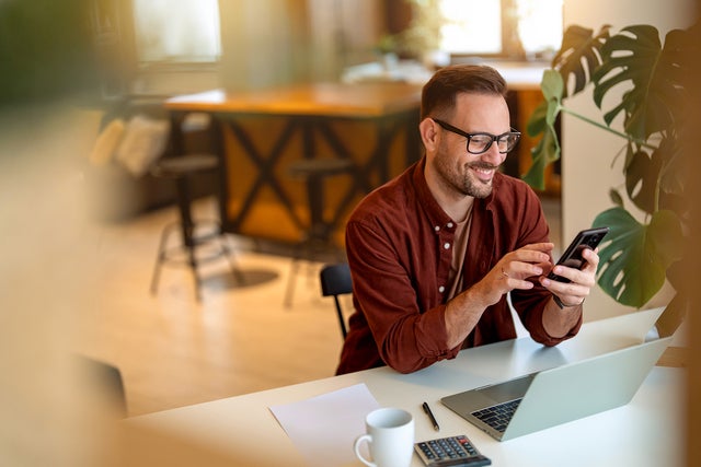 The picture shows a young man sitting at a white desk in his home, smiling and looking at his mobile phone and using the power metering service from ista. He is wearing a rust-brown corduroy shirt, a sand-brown shirt underneath and has black square glasses on. His hair is short and black and he has a short black beard. In front of him on the table is a laptop, next to it is a calculator, a ballpoint pen with a white sheet of paper and a white coffee cup. On the right side of the picture, directly next to the man, is a large monstera. The background is blurred, but a grey couch with brightly coloured cushions can be seen in the top left-hand corner and to the right of it is a orange and black wooden kitchen counter with two black bar stools.