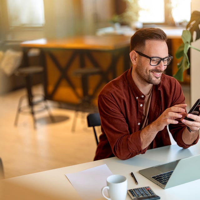 The picture shows a young man sitting at a white desk in his home, smiling and looking at his mobile phone and using the power metering service from ista. He is wearing a rust-brown corduroy shirt, a sand-brown shirt underneath and has black square glasses on. His hair is short and black and he has a short black beard. In front of him on the table is a laptop, next to it is a calculator, a ballpoint pen with a white sheet of paper and a white coffee cup. On the right side of the picture, directly next to the man, is a large monstera. The background is blurred, but a grey couch with brightly coloured cushions can be seen in the top left-hand corner and to the right of it is a orange and black wooden kitchen counter with two black bar stools.