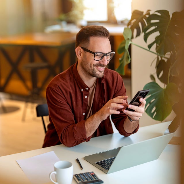 The picture shows a young man sitting at a white desk in his home, smiling and looking at his mobile phone and using the power metering service from ista. He is wearing a rust-brown corduroy shirt, a sand-brown shirt underneath and has black square glasses on. His hair is short and black and he has a short black beard. In front of him on the table is a laptop, next to it is a calculator, a ballpoint pen with a white sheet of paper and a white coffee cup. On the right side of the picture, directly next to the man, is a large monstera. The background is blurred, but a grey couch with brightly coloured cushions can be seen in the top left-hand corner and to the right of it is a orange and black wooden kitchen counter with two black bar stools.