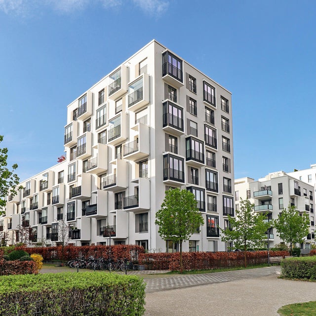 The picture shows a corner of a housing estate with white modern apartment buildings stretching from left to right across the picture. The buildings are characterised by an unusual architecture, as they alternately have protruding cubes with either large windows throughout the whole cube or set in balconies. The skyblue sky is filled with a few white clouds on the right side of the picture and in the foreground are red hedges and small trees that have been planted around the buildings, marking the property of the apartment buildings. There is a paved pedestrian path along the hedges and this is also bordered by another green hedge. The pedestrian path runs in a half circle in the bottom right of the picture, making a way through the grass that is planted there. 