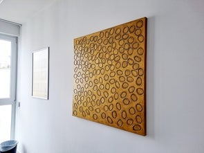 Ein Kunstwerk an einer weißen Wand. Das Gemälde hat eine goldene Hintergrundfarbe und besteht aus vielen ovalen Formen, die zufällig über die Leinwand verteilt sind. Auf der linken Seite des Bildes ist ein Fenster zu sehen.