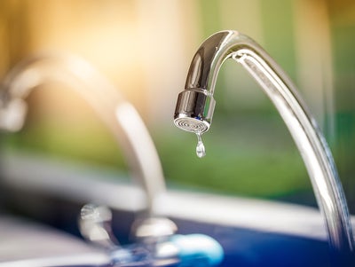 The picture shows a tap. The tap is made of shiny metal and has a curved shape that points downwards. At the end of the tap, where the water normally comes out, hangs a single drop of water that is about to fall. Another tap can be seen blurred in the background to the left of it. The entire background of the picture is blurred and shows a mixture of green and blue colours that could be reminiscent of an outdoor area or a garden. The light in the picture is warm and appears to be coming from the top left, casting a soft, golden glow on the tap. This creates a feeling of freshness and clarity, as if it is a sunny morning. In the foreground of the image, below the tap, there is a blurred area which may represent a sink or basin.