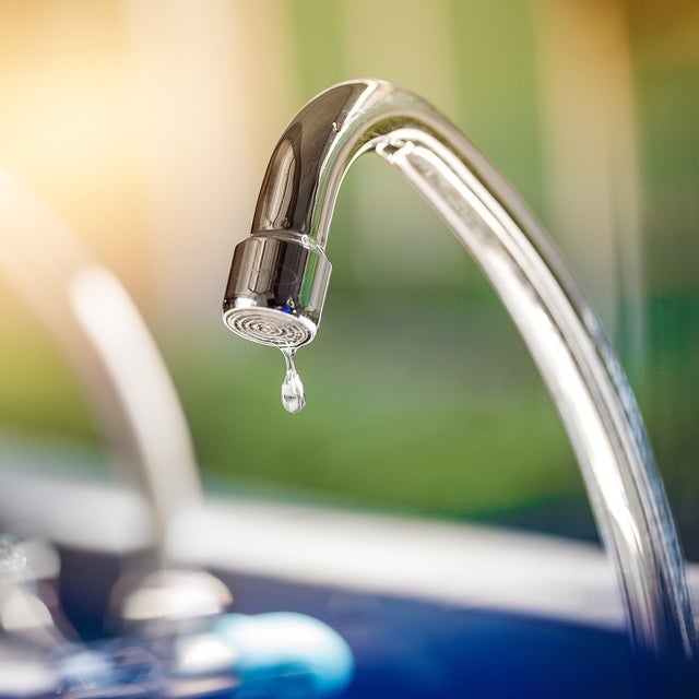 The picture shows a tap. The tap is made of shiny metal and has a curved shape that points downwards. At the end of the tap, where the water normally comes out, hangs a single drop of water that is about to fall. Another tap can be seen blurred in the background to the left of it. The entire background of the picture is blurred and shows a mixture of green and blue colours that could be reminiscent of an outdoor area or a garden. The light in the picture is warm and appears to be coming from the top left, casting a soft, golden glow on the tap. This creates a feeling of freshness and clarity, as if it is a sunny morning. In the foreground of the image, below the tap, there is a blurred area which may represent a sink or basin.