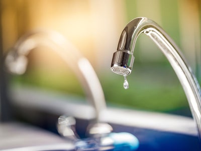 The picture shows a tap. The tap is made of shiny metal and has a curved shape that points downwards. At the end of the tap, where the water normally comes out, hangs a single drop of water that is about to fall. Another tap can be seen blurred in the background to the left of it. The entire background of the picture is blurred and shows a mixture of green and blue colours that could be reminiscent of an outdoor area or a garden. The light in the picture is warm and appears to be coming from the top left, casting a soft, golden glow on the tap. This creates a feeling of freshness and clarity, as if it is a sunny morning. In the foreground of the image, below the tap, there is a blurred area which may represent a sink or basin.