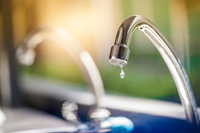 The picture shows a tap. The tap is made of shiny metal and has a curved shape that points downwards. At the end of the tap, where the water normally comes out, hangs a single drop of water that is about to fall. Another tap can be seen blurred in the background to the left of it. The entire background of the picture is blurred and shows a mixture of green and blue colours that could be reminiscent of an outdoor area or a garden. The light in the picture is warm and appears to be coming from the top left, casting a soft, golden glow on the tap. This creates a feeling of freshness and clarity, as if it is a sunny morning. In the foreground of the image, below the tap, there is a blurred area which may represent a sink or basin.