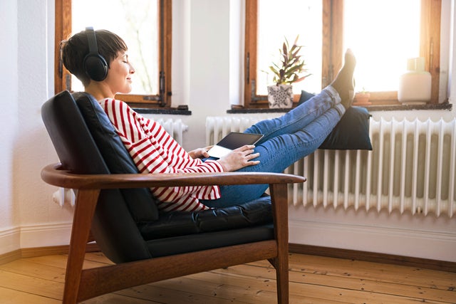 The picture shows a woman sitting comfortably on an armchair with wooden armrests and a black leather seat in front of a window. She has short brown hair and is wearing a white and red striped jumper and blue jeans. She is holding a tablet on her lap and listening to music through over-ear headphones. Her feet are propped up on a cushion resting on a radiator in front of the window. The floor of the room is made of light-coloured wood. On the windowsill, there is a flower pot with a green plant on the left and an empty white vase on the right.